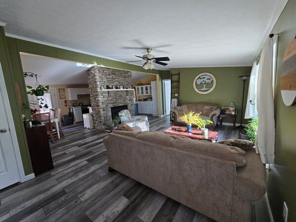 A living room with a stone fireplace, a ceiling fan, and a dining area in the background.