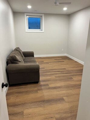 A room with a brown sofa, wooden flooring, a ceiling fan, and a window with blinds.