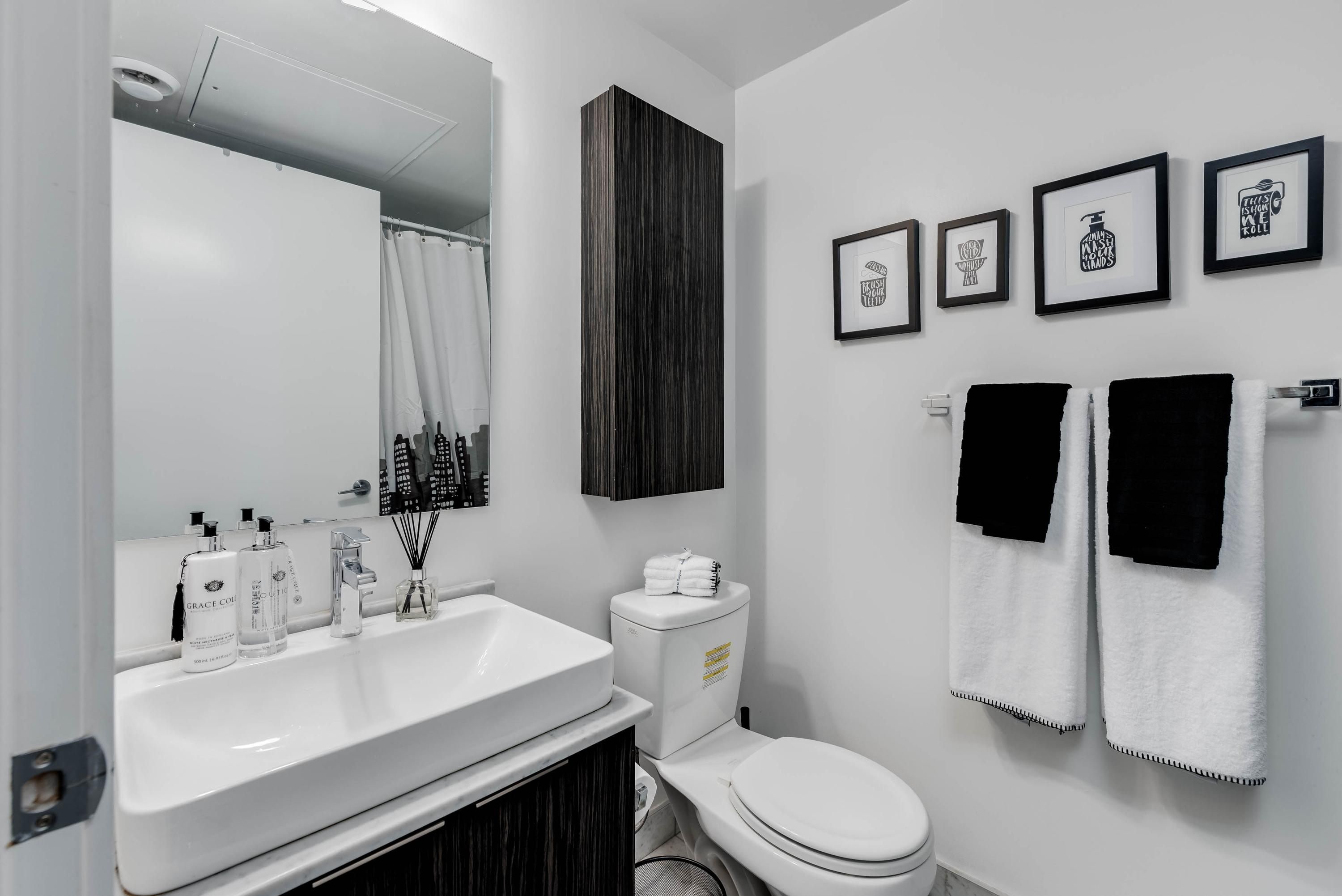 A modern bathroom with a white sink, toilet, and a large mirror. There are framed pictures on the wall and two towels hanging on a rack.