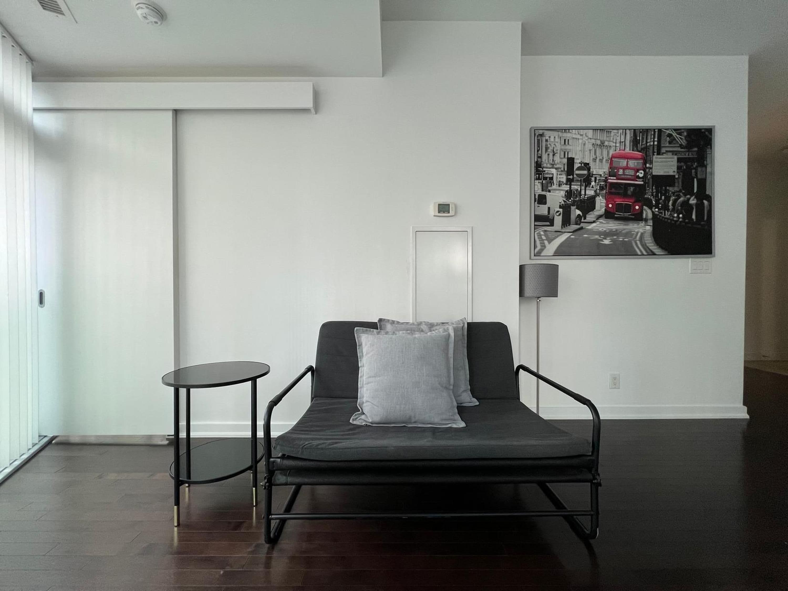 A modern room with a black sofa, a small round table, and a black lamp. A black and white photo of a street scene is on the wall.