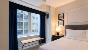Ein Hotelzimmer mit Bett, Nachttisch mit Lampe, einer Bank, einem Fenster mit Vorhängen und gerahmten Bildern an der Wand.
