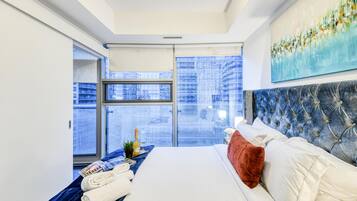 A hotel room with a large bed, a window with a view of buildings, a painting on the wall, and a small table with a bottle and glasses.