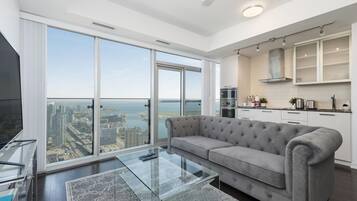 A modern living room with a large glass coffee table, a grey sofa, and a city view through floor-to-ceiling windows.