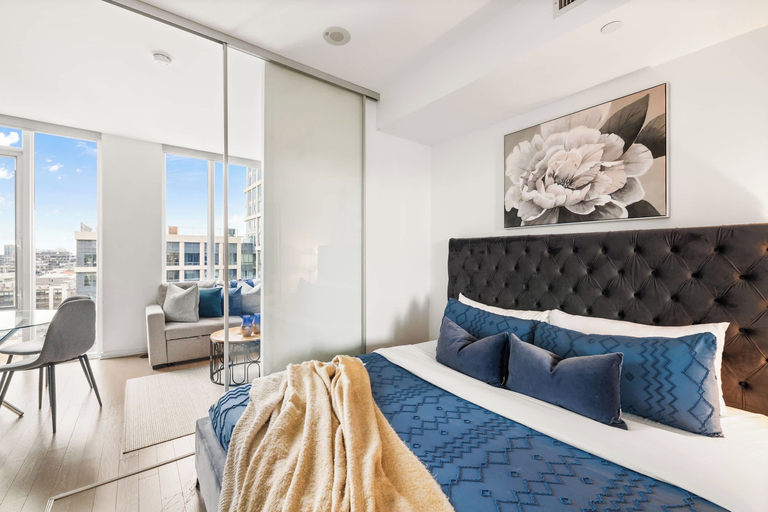 A modern bedroom with a large bed, a city view, and a dining area with a table and chairs.