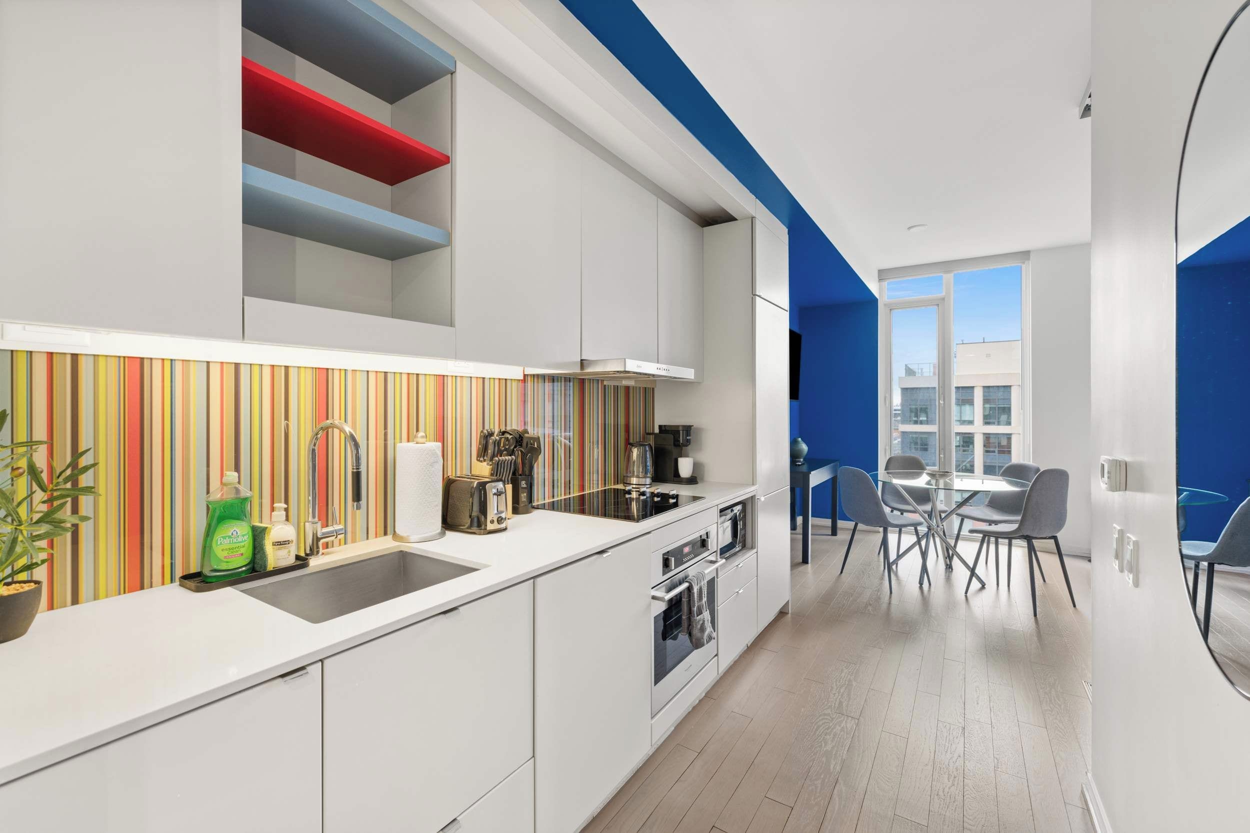 A modern kitchen with white cabinets, a striped backsplash, and a dining area with a blue accent wall and a glass table.