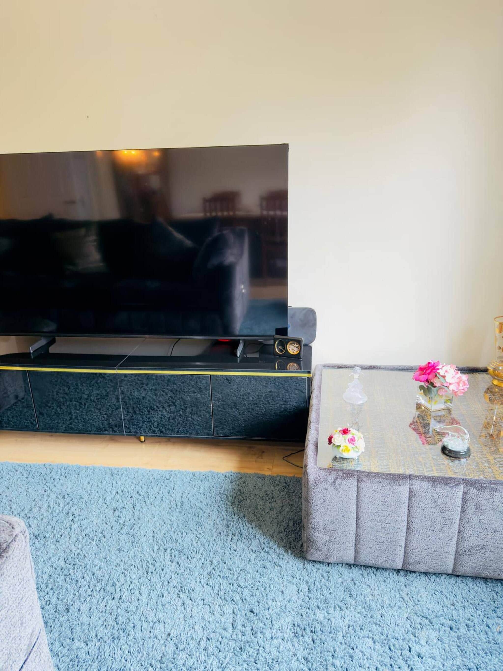 A flat-screen TV on a stand, a glass coffee table with flowers, and a blue carpet.
