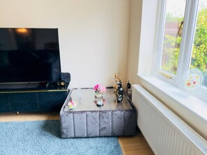 A living room with a flat-screen TV, a glass coffee table with decorative items, a sofa, and a window with a view of greenery. - More Photos to Come in few Weeks (Norwich)