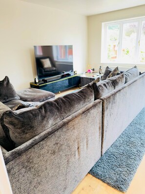 A modern living room with a large sectional sofa, a flat-screen TV, and a window with natural light. - More Photos to Come in few Weeks (Norwich)
