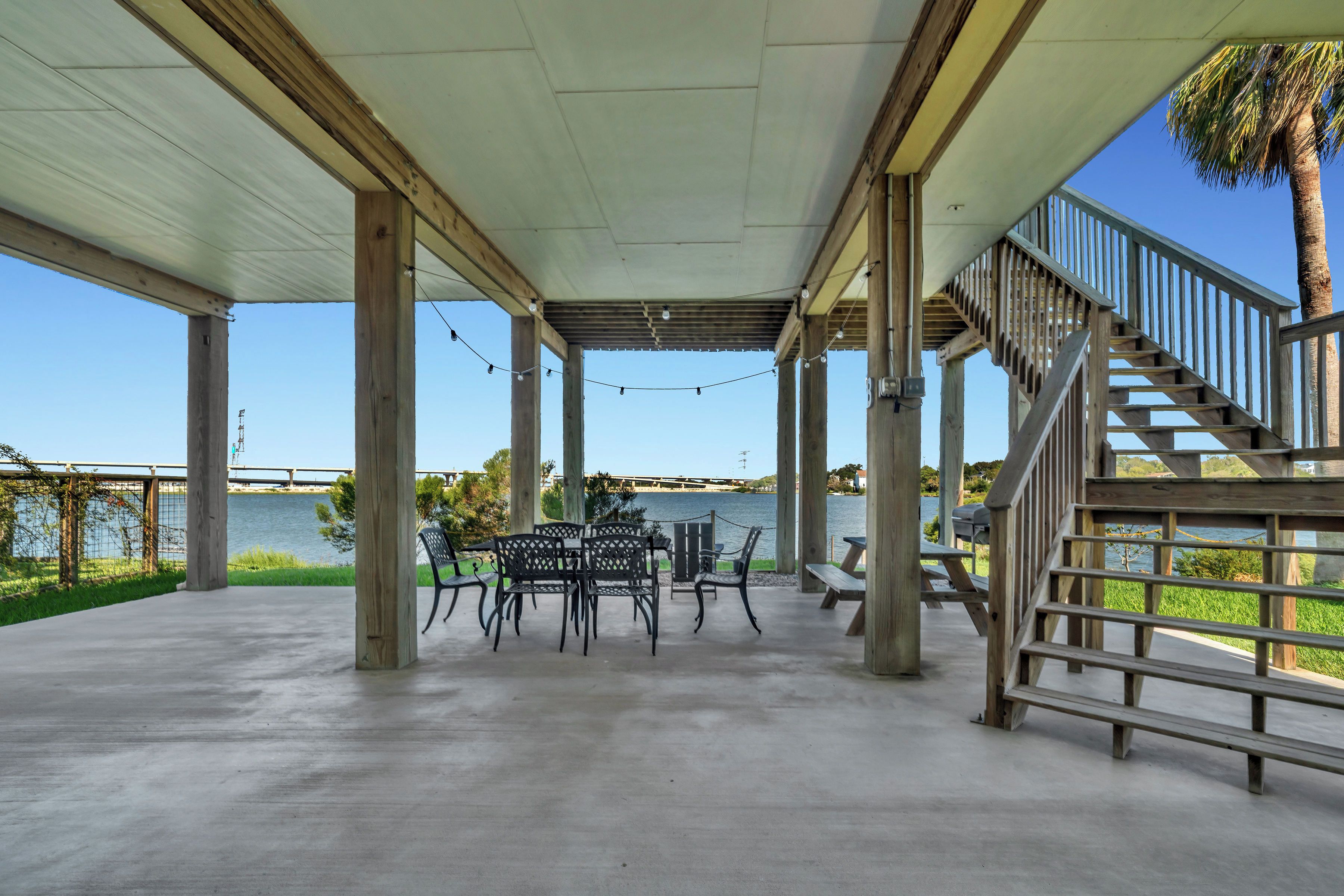 An outdoor covered patio with a view of a body of water, featuring a table and chairs, a staircase, and a railing.
