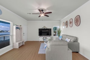 Una sala de estar con ventilador de techo, televisor de pantalla plana, sofá gris y vista al agua a través de un ventanal.