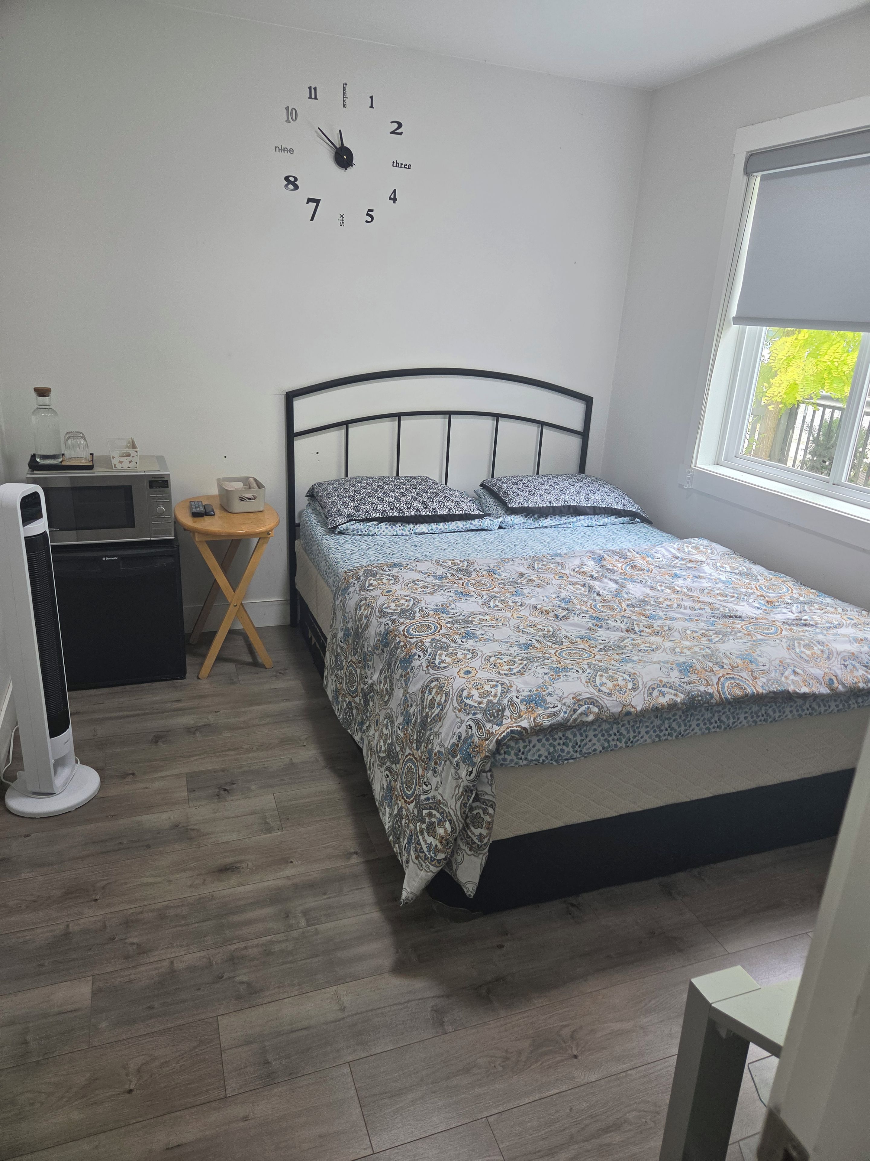 A bedroom with a bed, a wall clock, a bedside table with a lamp, and a window.