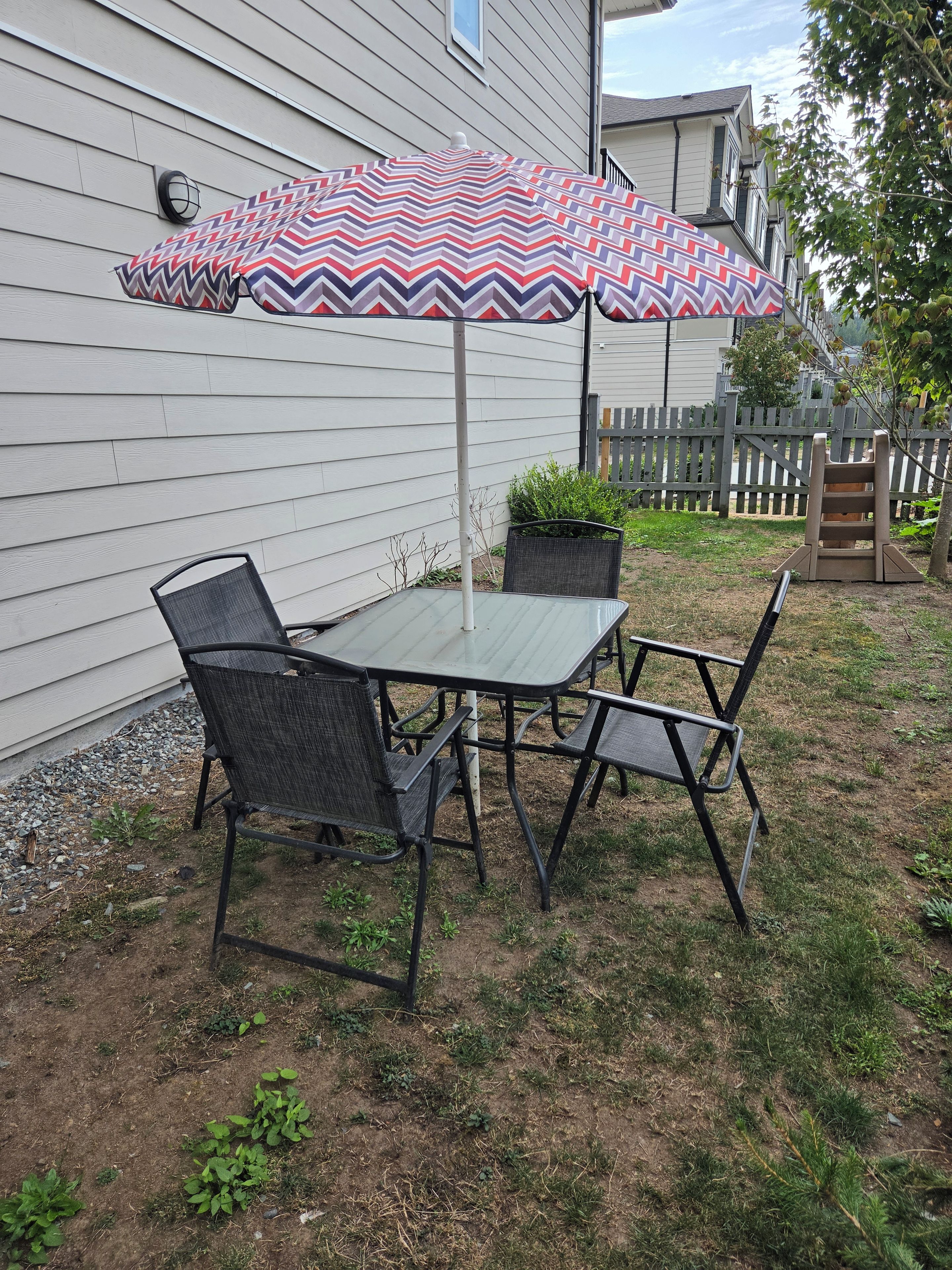 An outdoor patio with a table and chairs, a patterned umbrella, and a wooden fence in the background.