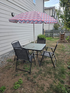 An outdoor patio with a table and chairs, a patterned umbrella, and a wooden fence in the background. - Serene 1-bedroom townhome in welcoming Surrey neighborhood (Surrey)