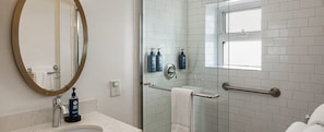 A bathroom with a round mirror, a sink with a marble countertop, a glass shower enclosure, and a window. - Celebrate NYE in San Francisco — Luxury 1BR Hotel Suite (San Francisco)