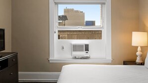 Ein Hotelzimmer mit einem Bett, einem Fernseher auf einer Kommode, einem Fenster mit Blick auf Gebäude und einer Klimaanlage.