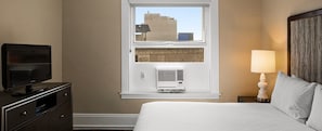 A hotel room with a bed, a television on a dresser, a window with a view of buildings, and an air conditioning unit. - Celebrate NYE in San Francisco — Luxury 1BR Hotel Suite (San Francisco)