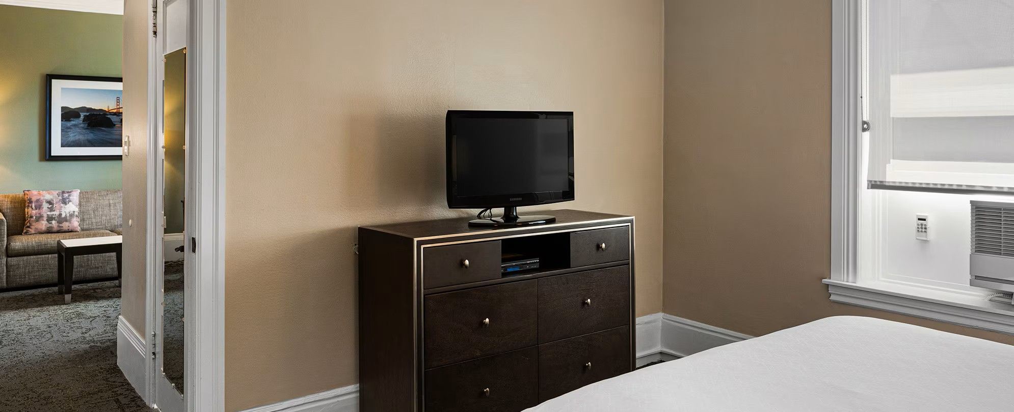 A hotel room with a bed, a dresser, a television on top, and a window with blinds.