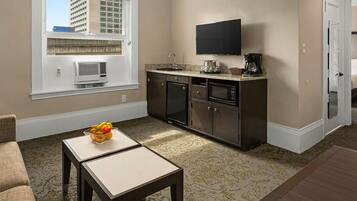 A hotel room with a kitchenette, a flat-screen TV, a dining area with a table and chairs, and a view of the cityscape through the window.