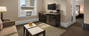 A hotel room with a kitchenette, a flat-screen TV, a dining area with a table and chairs, and a view of the cityscape through the window.