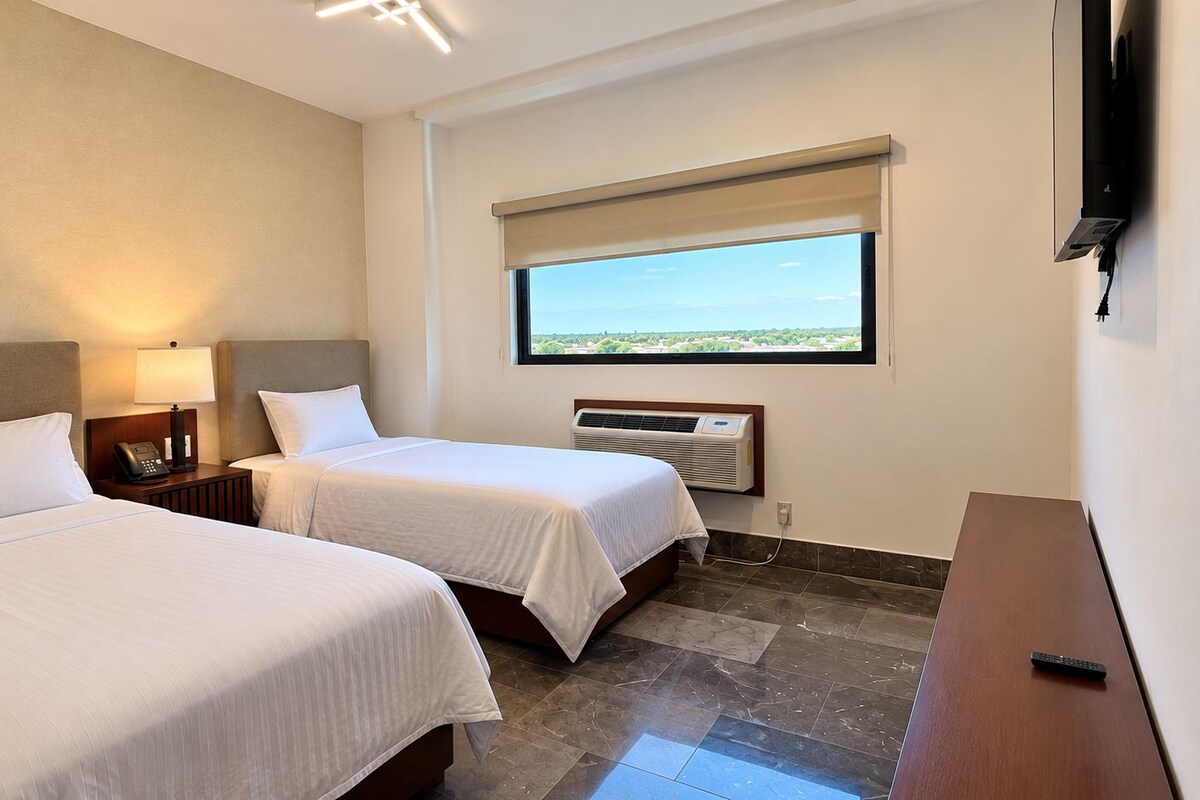 Habitación de hotel con dos camas, una mesita de noche con lámpara, televisión montada en la pared y ventana con vistas.