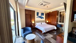 Habitación de hotel con una cama, una silla, una mesita, una mesita de noche y un cuadro en la pared.