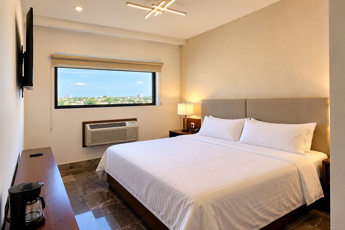 Una habitación de hotel con cama, escritorio, silla, televisor, ventana y aire acondicionado de pared.