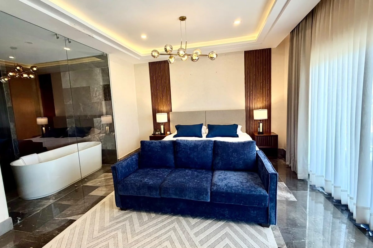 Ein modernes Hotelzimmer mit einem blauen Sofa, einer Duschkabine aus Glas und einem großen Fenster mit Vorhängen.