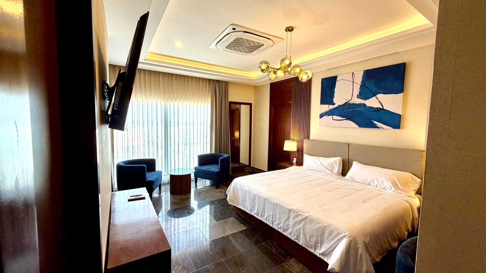 A hotel room with a bed, two armchairs, a small table, a TV, and a painting on the wall.