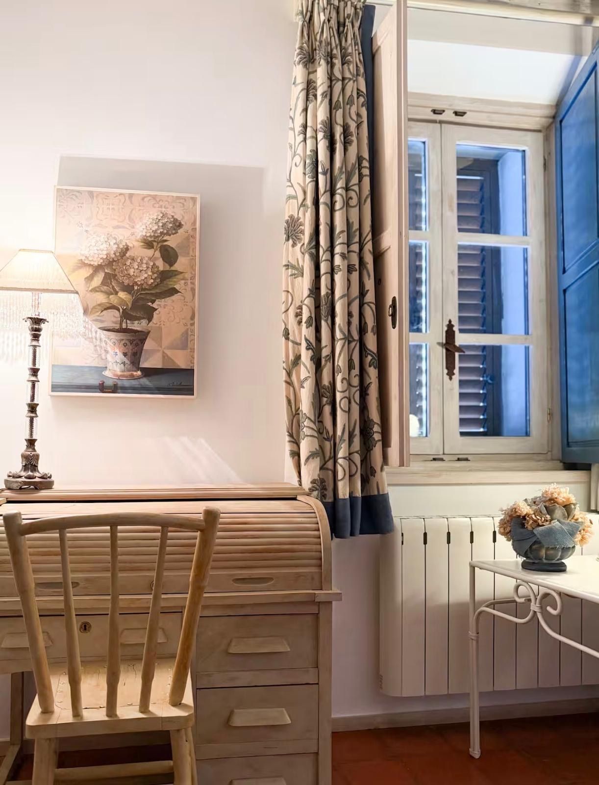 Una habitación con una silla de madera, una mesa, una lámpara, una ventana con contraventanas azules, una cortina con estampado floral y un cuadro de flores en la pared.