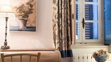 Una habitación con una silla de madera, una mesa, una lámpara, una ventana con contraventanas azules, una cortina con estampado floral y un cuadro de flores en la pared.