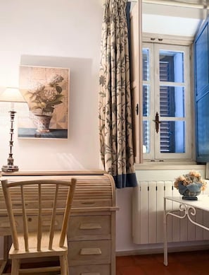 Een kamer met een houten stoel, een tafel, een lamp, een raam met blauwe luiken, een gordijn met een bloemmotief en een bloemen schilderij aan de muur.