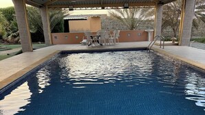 Una piscina con solárium azulejado, zona de estar cubierta y pared de piedra al fondo.
