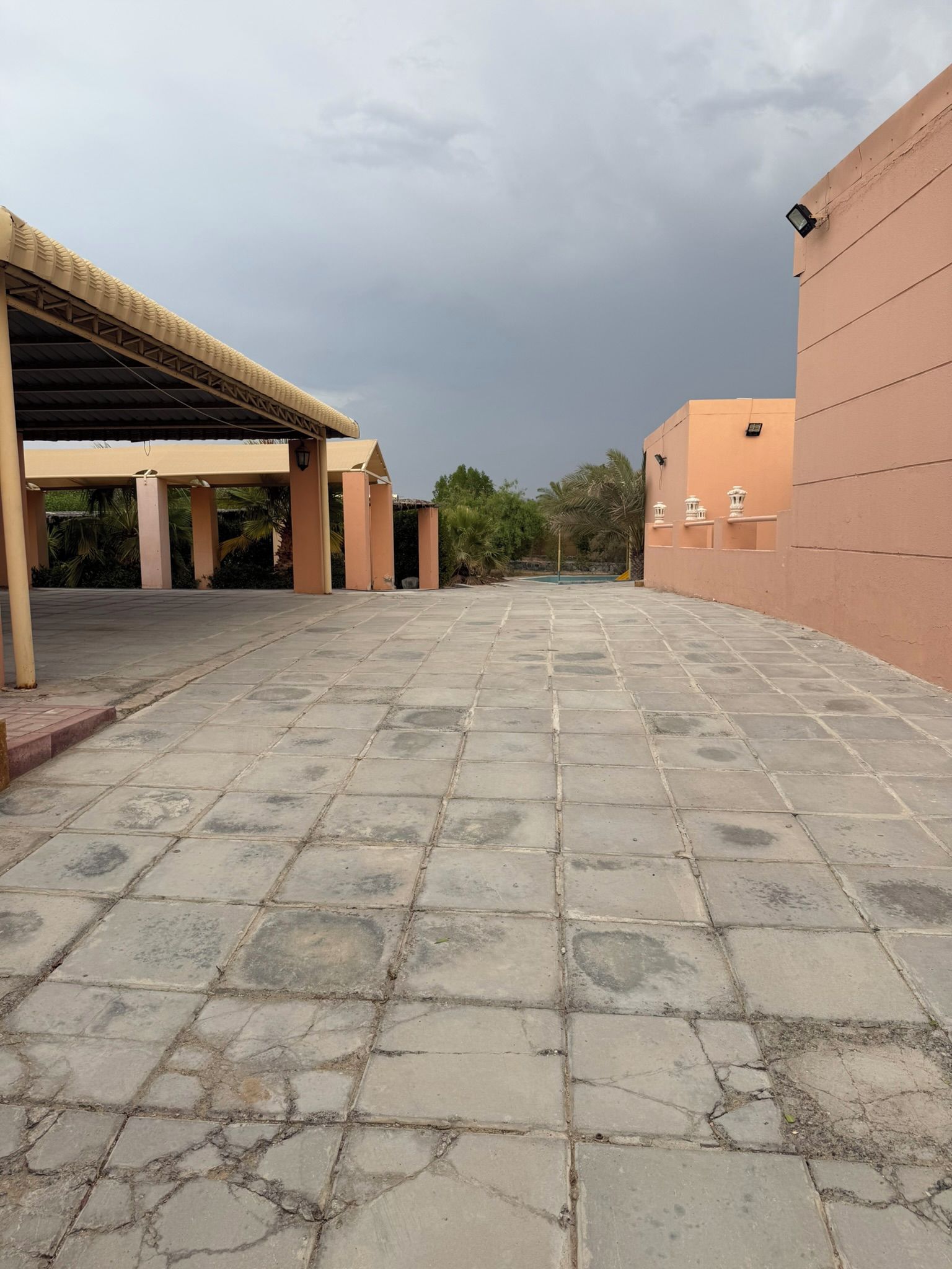 Un patio empedrado con un corredor cubierto, palmeras y un edificio de fachada color beige.