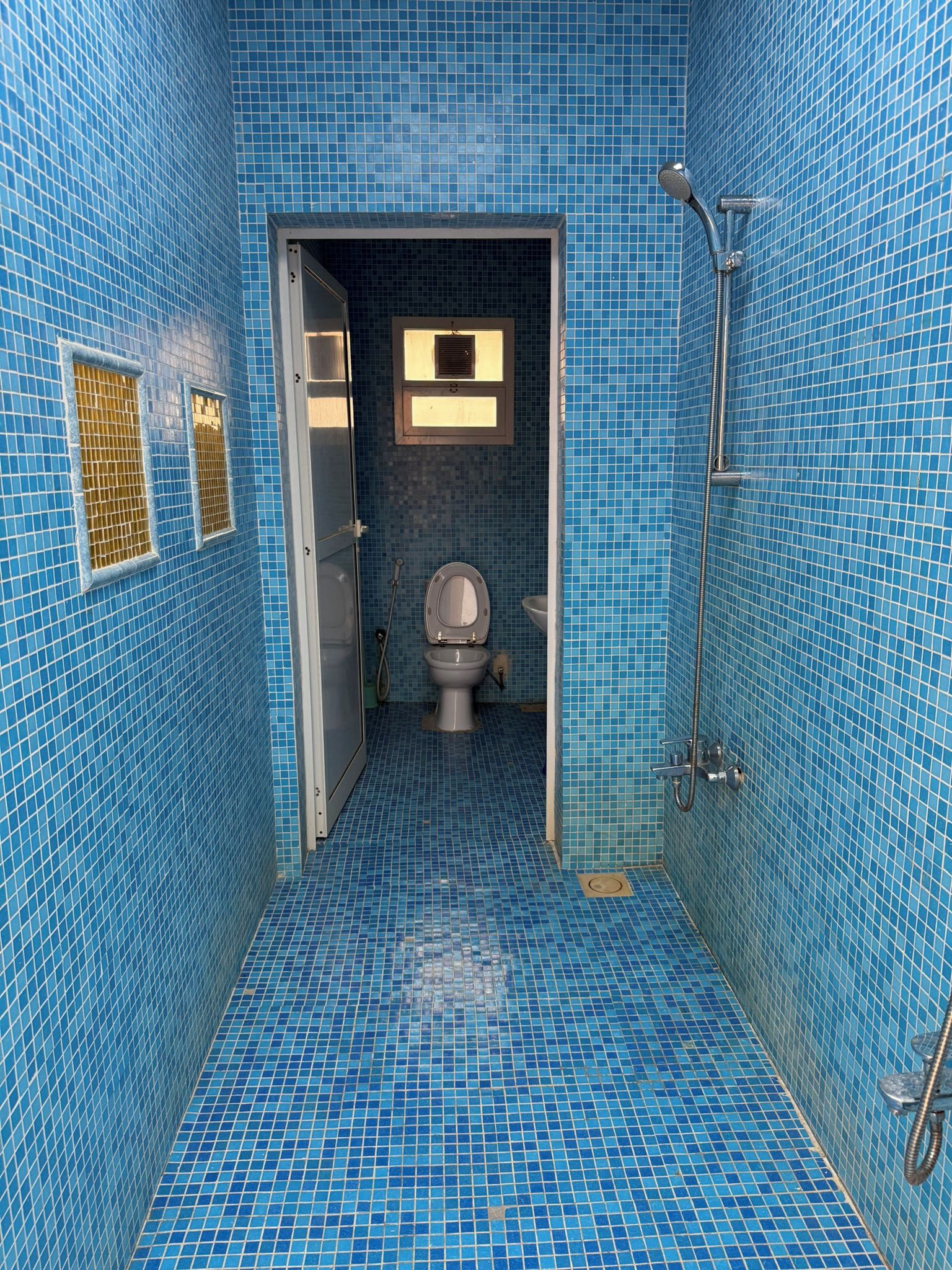 Een badkamer met blauw betegelde wanden en vloer, een toilet, een douche en een raam met zonwering.