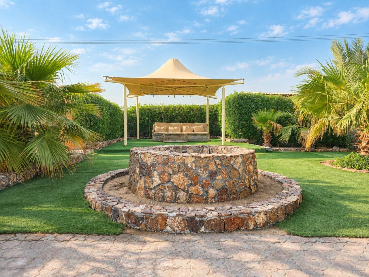 A circular stone fire pit surrounded by a stone border, with a canopy overhead and a bench underneath.