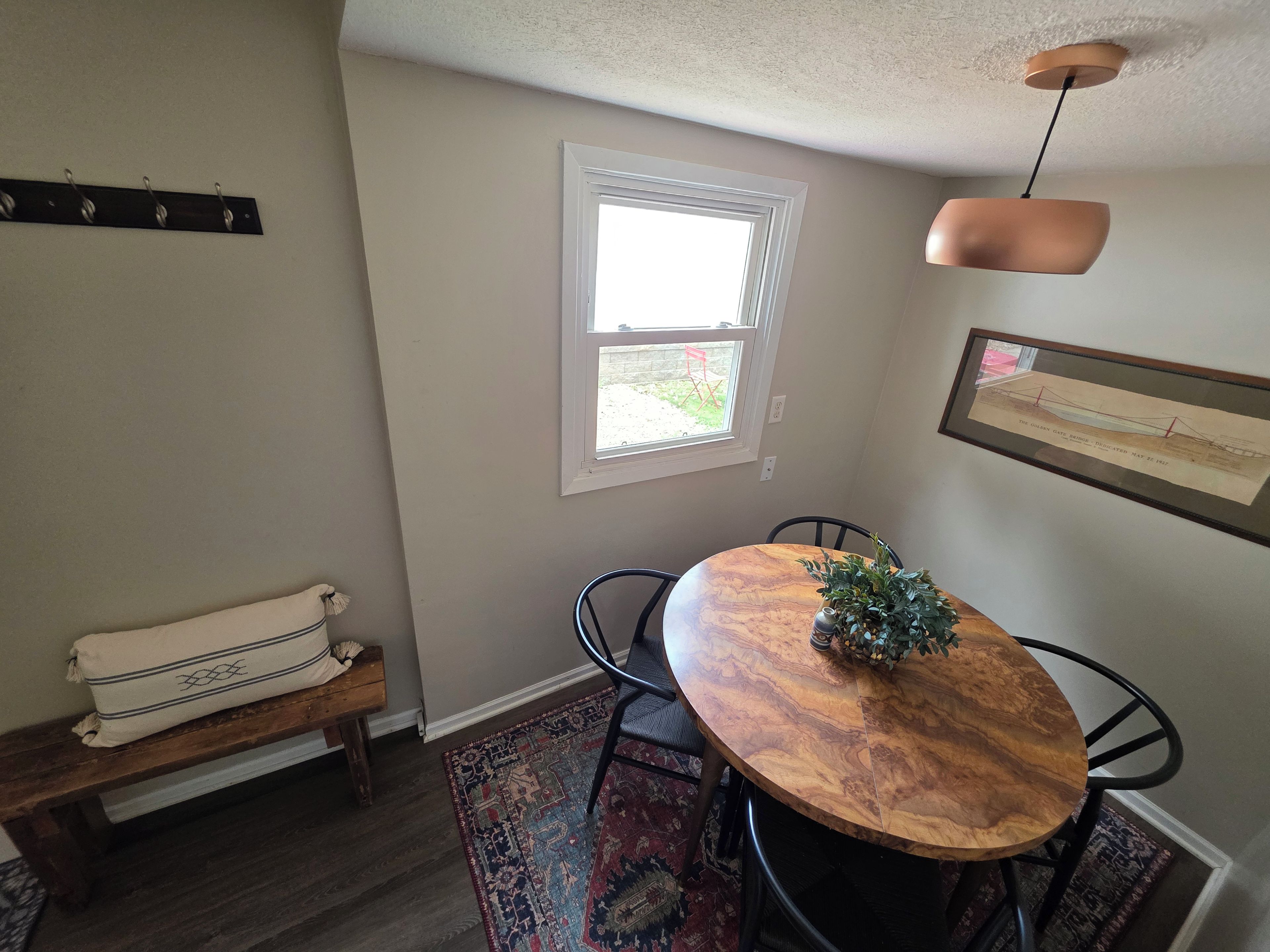 A dining area with a wooden table, black chairs, a bench, a hanging light fixture, a window, and a framed picture on the wall.