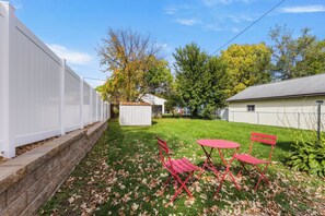 A backyard with a white fence, a small shed, a red table and chairs, and a lawn with scattered leaves.