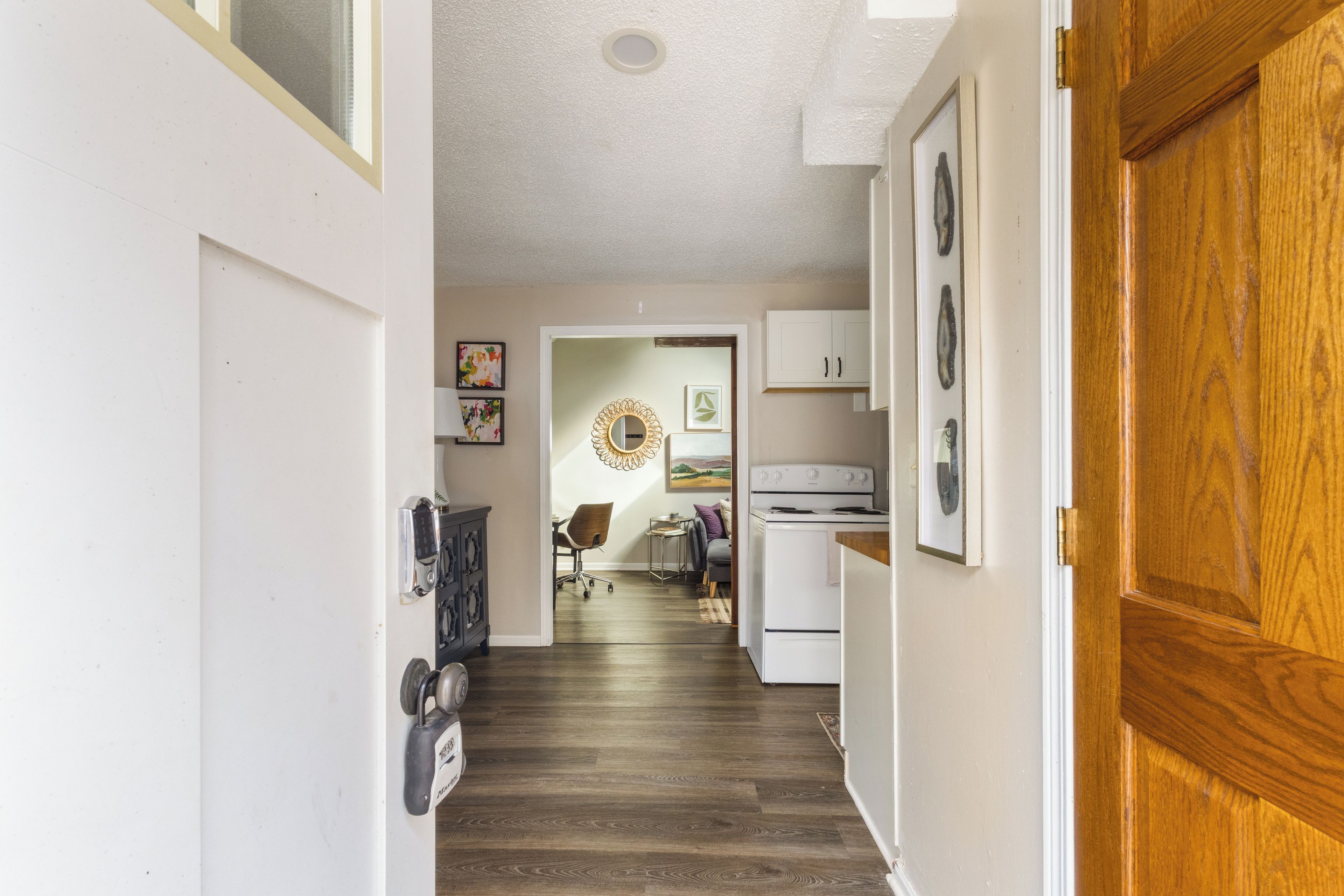 A hallway with a wooden floor, a white door, and a kitchen area with a stove and cabinets.