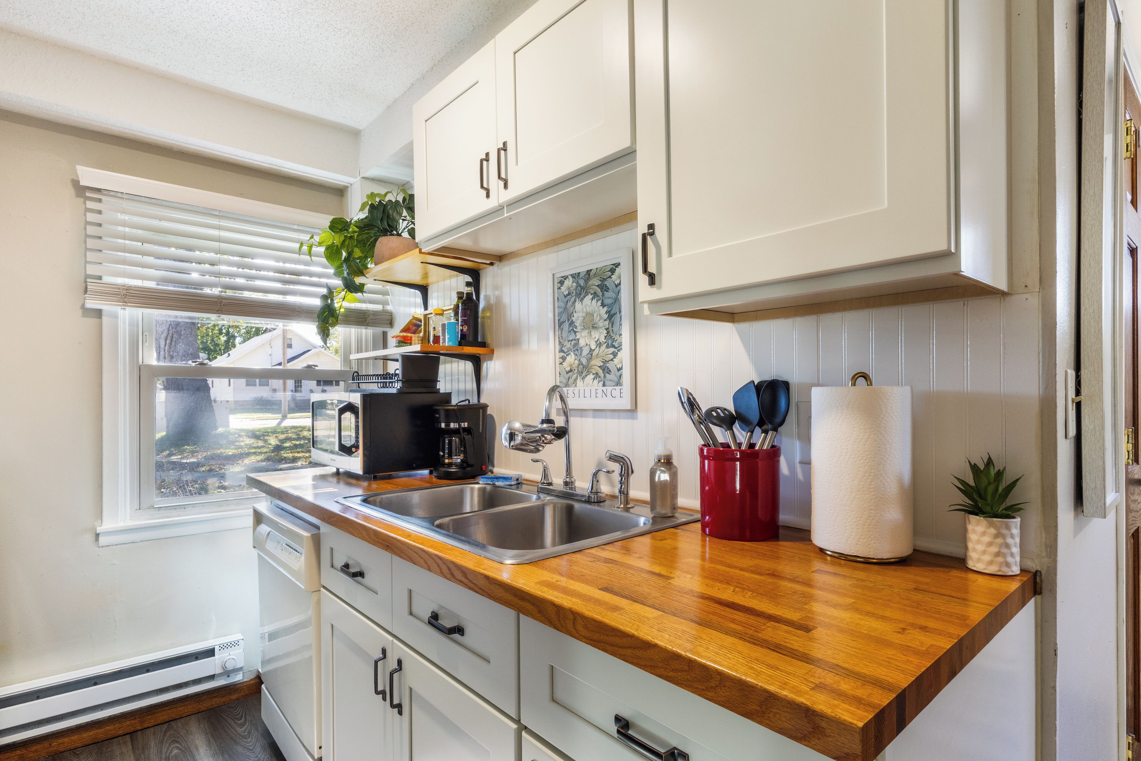 Cocina con armarios blancos, encimera de madera, un lavamanos, cafetera y ventana con persianas.