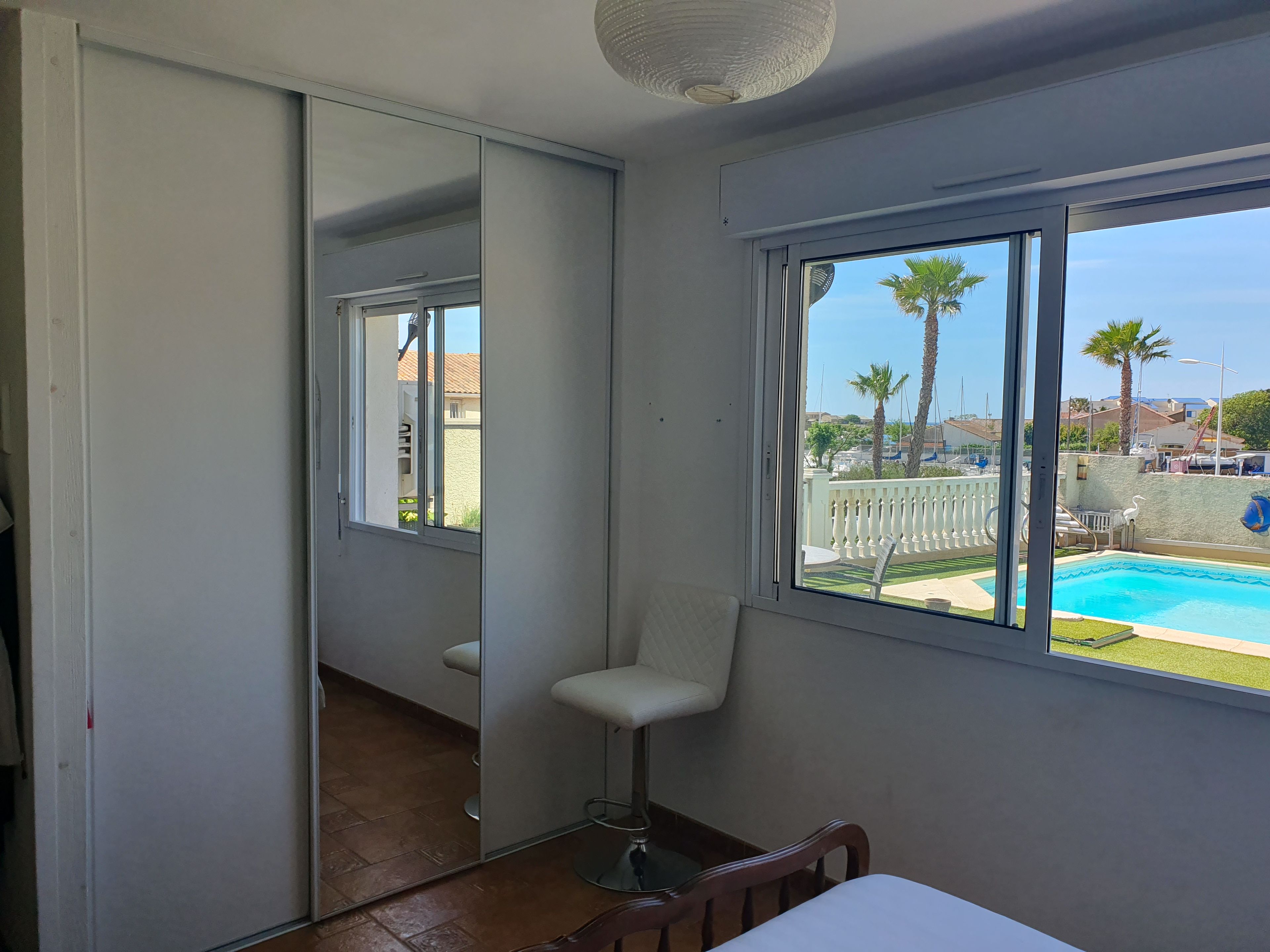 A room with a large window, a white chair, a small table, and a mirror reflecting the outside view.