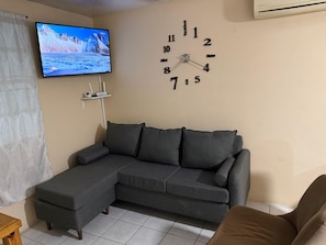 Ein Wohnzimmer mit einem grauen Ecksofa, einem flachbildfernseher an der Wand und einer Wanduhr.