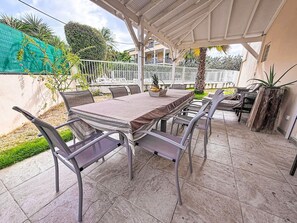 Eine Terrasse im Freien mit einem überdachten Tisch und Stühlen, umgeben von einem weißen Zaun und viel Grün.