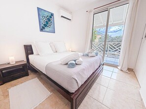Ein Schlafzimmer mit einem Bett, einem Nachttisch, einem Gemälde, einer Klimaanlage und Blick durch eine Schiebetür.