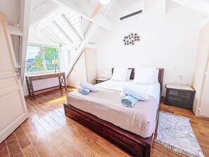 Quarto com cama de madeira, mesa de cabeceira, escrivaninha e vista para o exterior.