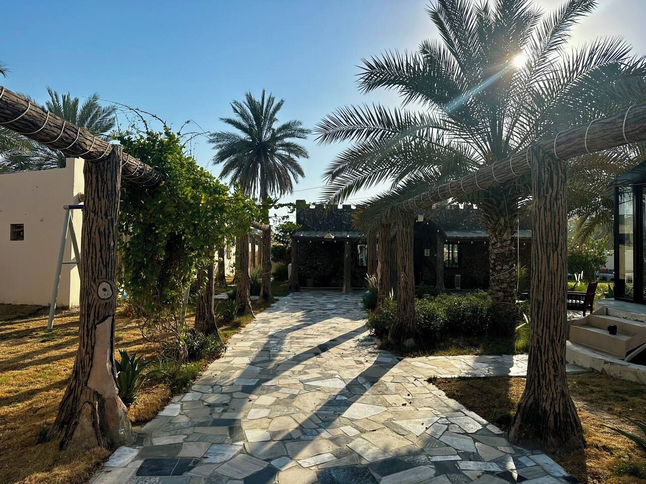 A stone pathway leading to a covered area with palm trees and a building in the background.