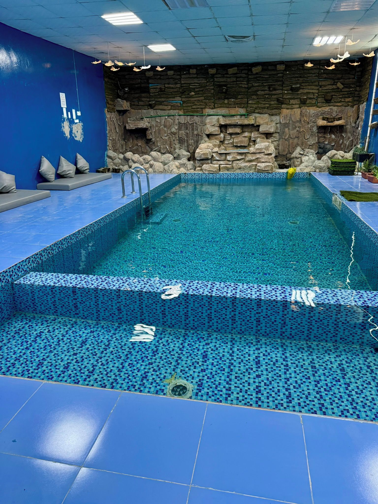 An indoor swimming pool with a stone wall backdrop, tiled flooring, and a blue color scheme.