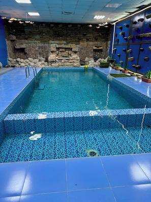 Ein rechteckiger, überdachter Schwimmteich mit blauen Mosaikfliesen, einer Wand aus Stein und Topfpflanzen.