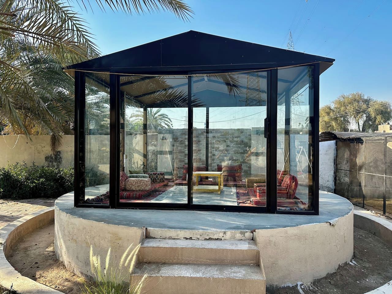 A glass-enclosed gazebo with a curved roof, a central seating area with cushions, and a concrete base.