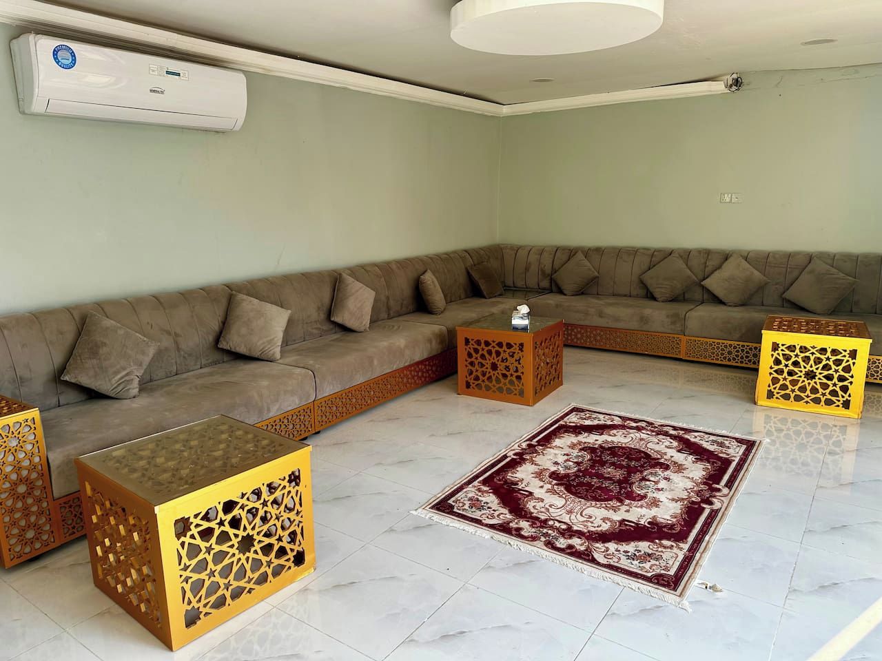 A living room with a sectional sofa, two decorative wooden side tables, a patterned rug, and an air conditioning unit on the wall.