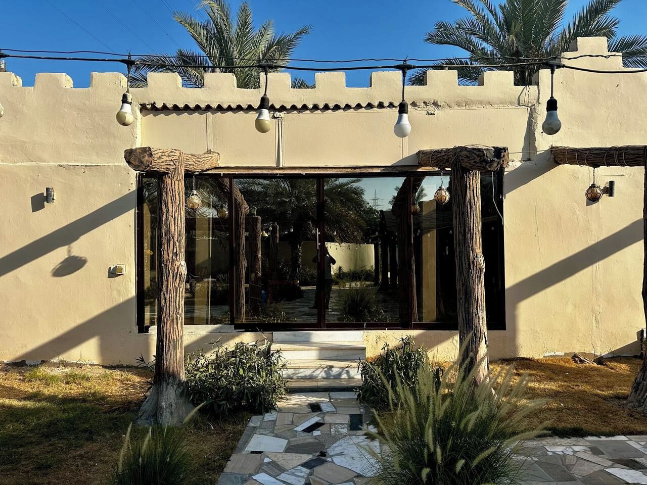 A building entrance with a stone pathway leading to a glass door, surrounded by palm trees and a clear sky.
